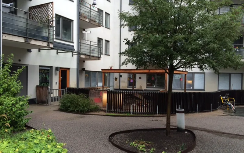 Förskolans entré med utemiljö och ett träd i förgrunden. Vitt flerbostadshus i bakgrunden.