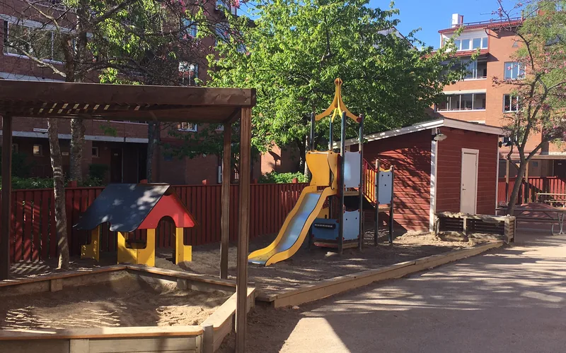Lekredskap och sandlåda på den soliga förskolegården, bostadshus i bakgrunden.