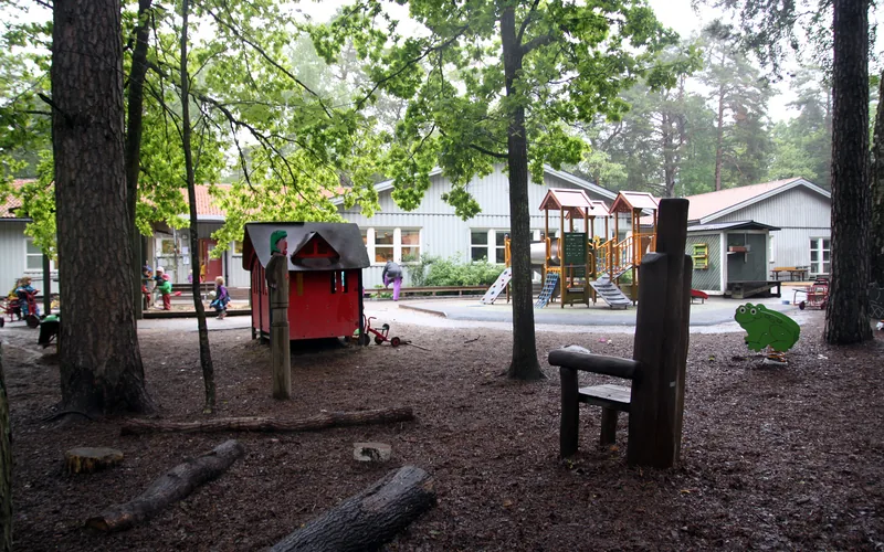Förskolegården med lekredskap, träd och stockar, förskolans hus i bakgrunden.