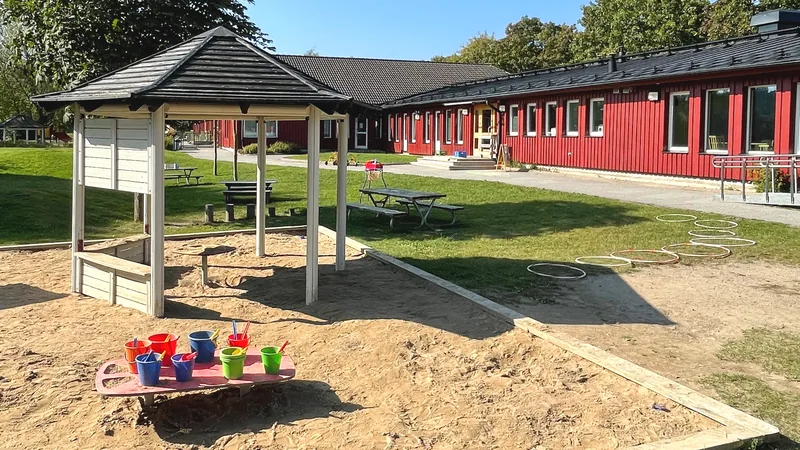En förskolegård med en stor sandlåda och ett lekbord med hinkar och spadar. Rockringar ligger utlagda och i bakgrunden syns picknickbord och en röd envåningsbyggnad,