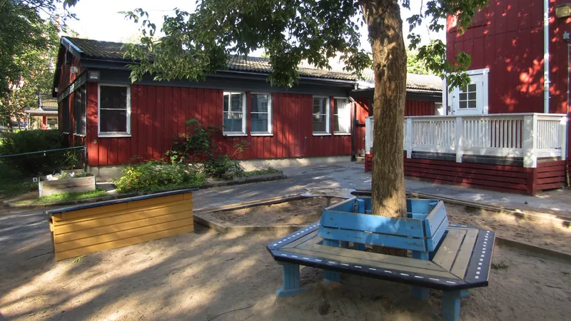 Röda träfasader och lekmiljö med sandlåda och träd i förgrunden.