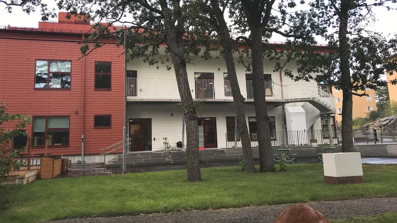 Husfasad med gräs och höga träd i förgrunden.