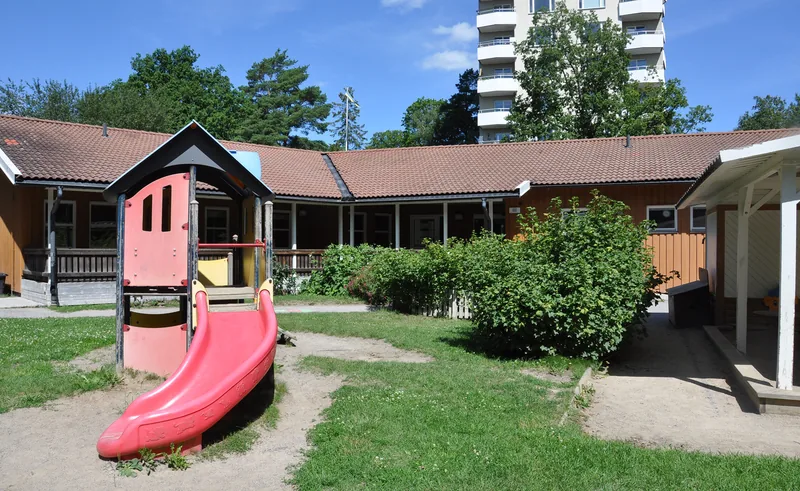 Ett gult trähus i ett plan med stor lekyta framför, sandlåda och lekredskap med rutschkana.