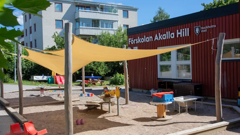 Förskolegård med träd, buskar, gräs, rutschkana, bord och stor sandlåda med solskydd och leksaker. I övrigt asfalt.