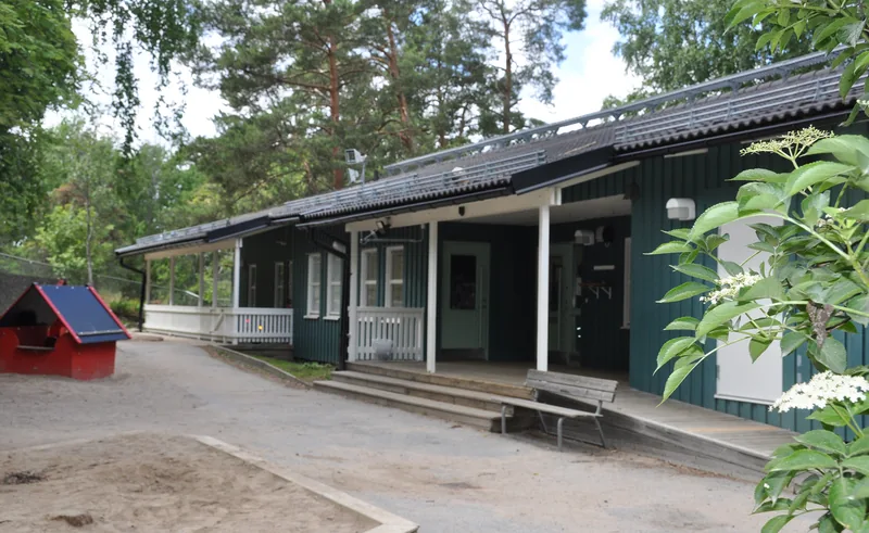En låg träbyggnad med stort skogsparti framför och en rutschkana.