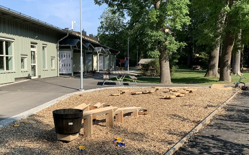 Förskola i bakgrunden med gård framför sig med lekhus, sandlåda, cyklar, bord att sitta vid och grönska.