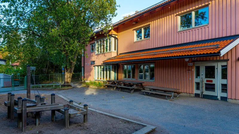 Förskolebyggnad i två plan med sittplatser och lekredskap utomhus.