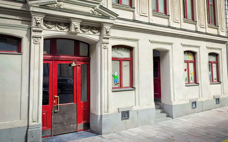Entré från gatan till förskolan Flaggskepet. Stenbyggnad med röd dörr och fönster som är smyckade med olika pynt. Foto.