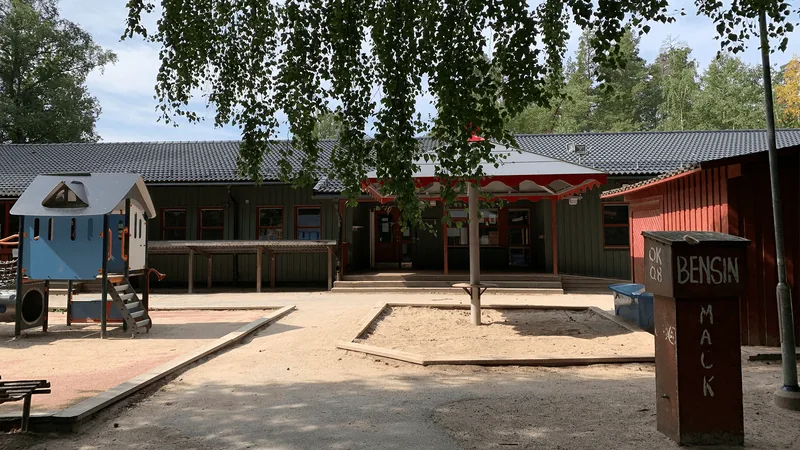 Ett enplanshus i trä med stor gård. På gården finns sandlådor, stora träd, rutschkana och bänkar.