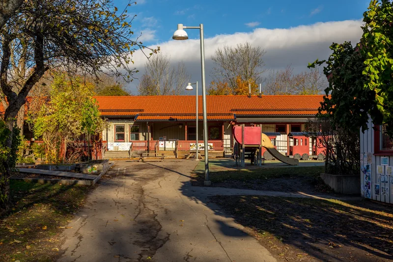 Byggnad med asfalt, buskar träd, klätterställning och lektåg på gården framför sig.