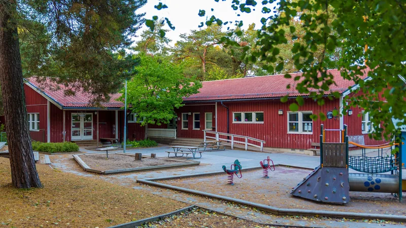 Röd träbyggnad inklämd bland grönskande träd. Lekplats i förgrunden, foto.