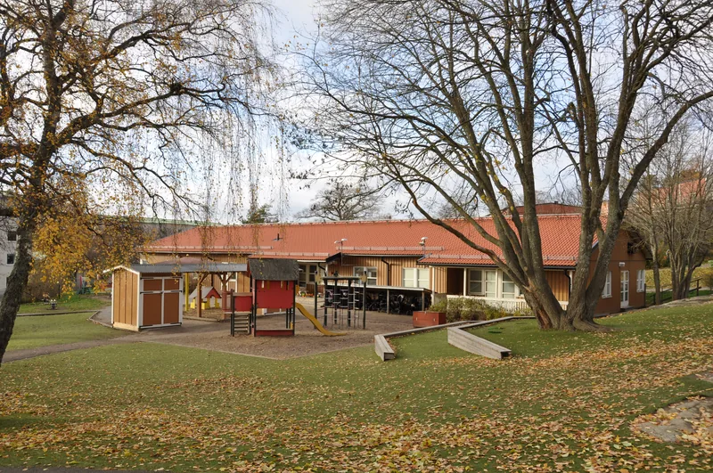 Låg förskolebyggnad med träpaneler, träd och en stor lekyta framför med många lekredskap.