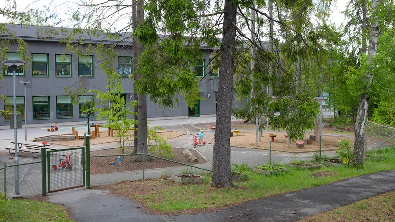 Förskolegård med träd, stubbar, stenar och sandlådor. I övrigt asfalt med bord, trehjulingar och andra leksaker.