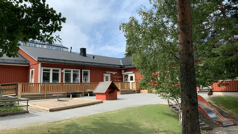 Ett enplanshus i trä, med stor gård i en backe. På gården finns sandlådor, stora träd, rutschkana och bänkar.