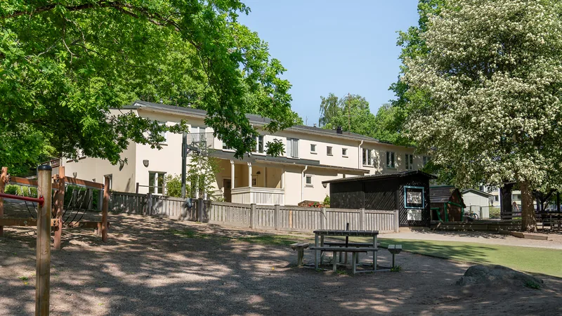 En förskolebyggnad och en gård med lekredskap och sittplatser runt grönska