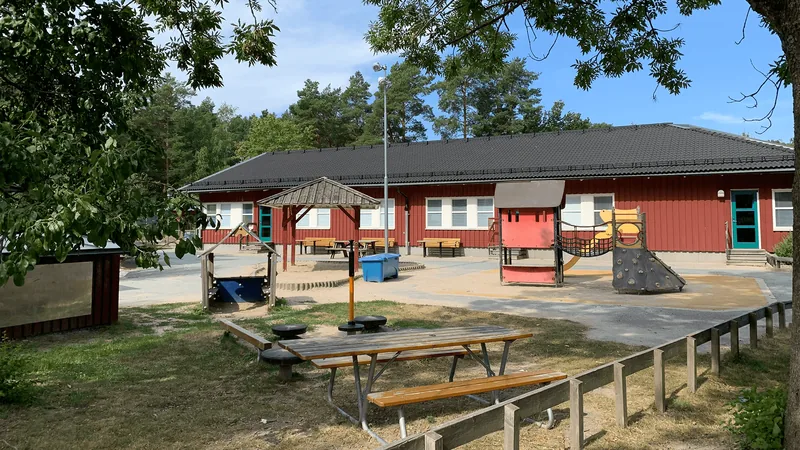 Stor gård med bänkar och lekplats. Ett enplanshus i trä i bakgrunden.