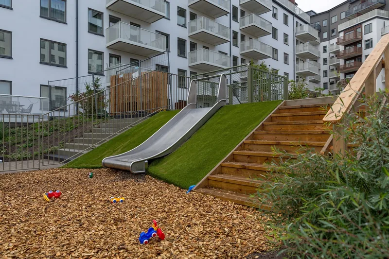 Rutschkana och buskar på gården med flerfamiljshus i bakgrunden.