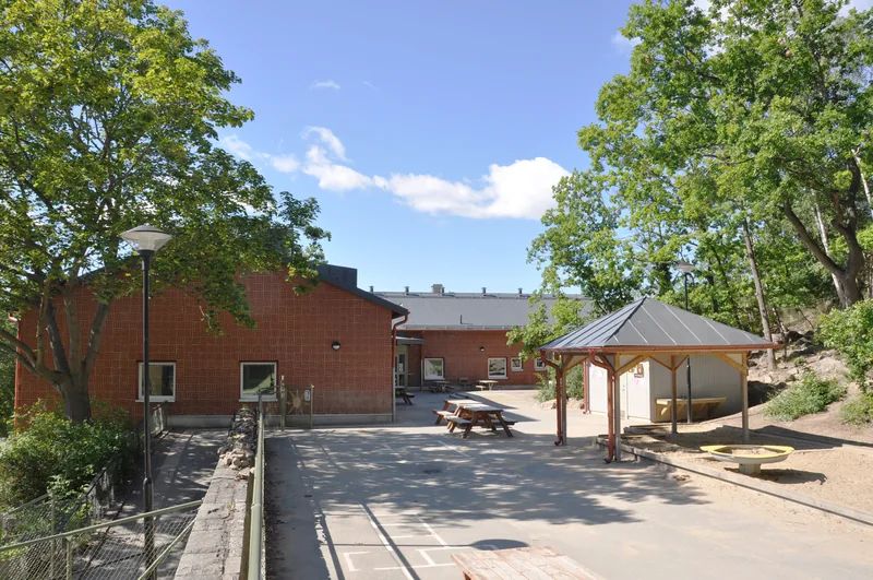 Låg tegelbyggnad med stor gård med grönskande träd, bänkar och pergola.