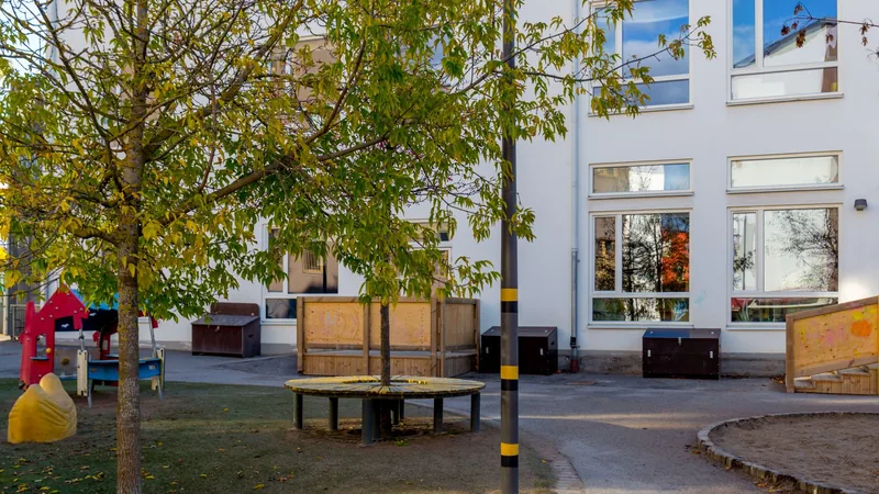 Utegård och lekplats syns framför vit förskola med stängsel.