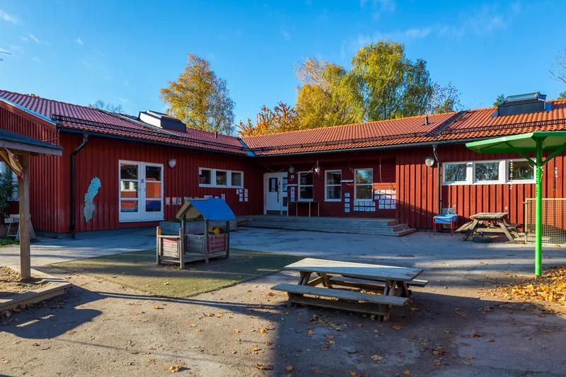 Byggnad med asfalt framför med sittplatser och lekobjekt samt träd i bakgrunden.