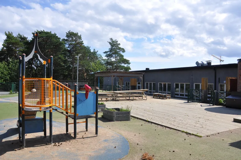 Ett enplanshus på en solig gård med staket runt om, på gården finns rutschbana, odlingslådor och sittbänkar.