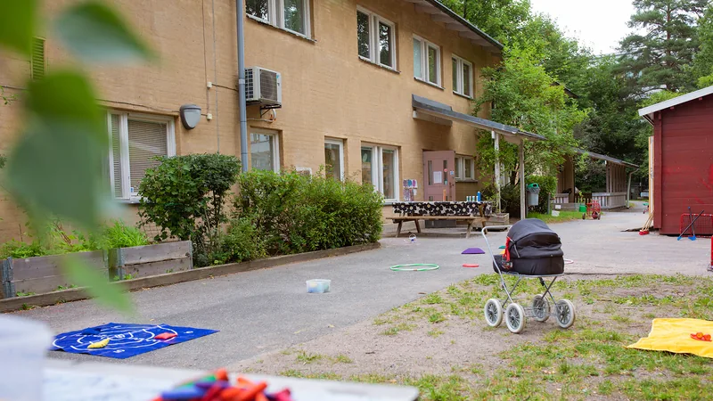 Förskolegård medträd, buskar, planteringar och gräsmatta. I övrigt asfalt med bord, trehjulingar, mål och bandyklubbor.