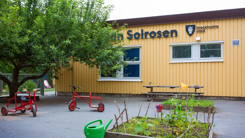 Förskolegård med träd, planteringar, gräsmatta och rutschkana. I övrigt asfalt med bord, trehjulingar och andra leksaker.