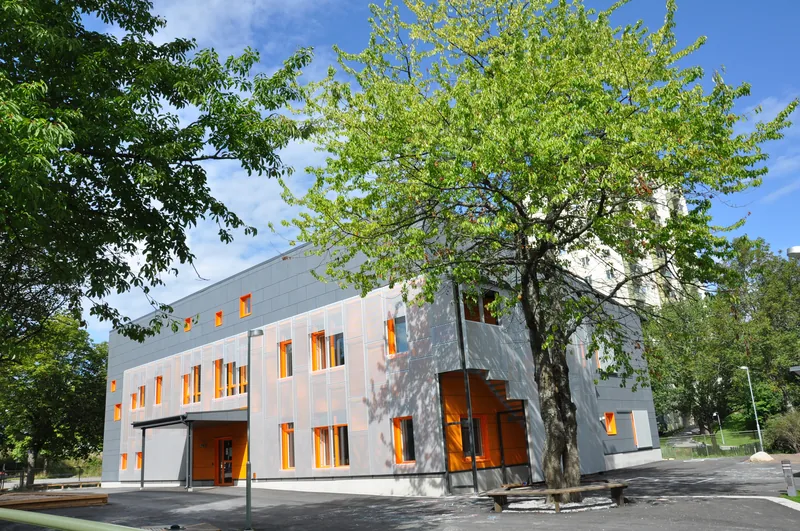 Modern tvåvåningsbyggnad, en solig gård och träd med spirande löv.