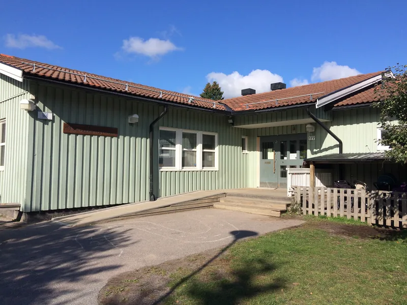 Solig huvudentré till ett grönt enplanshus i trä, med gräsmatta utanför.
