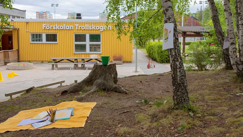 Förskolegård med träd, kuperad terräng och buskar. I övrigt asfalt med bord, trehjulingar och andra leksaker samt sandlåda.