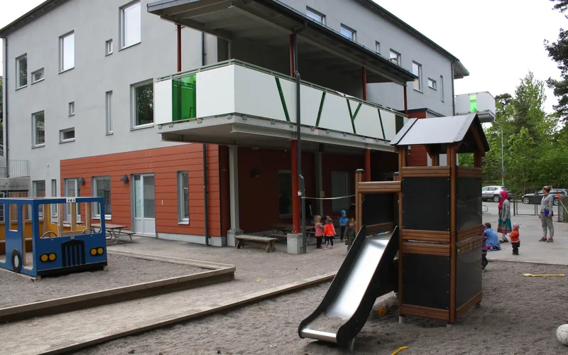 Byggnad med grå fasad, barn som leker. I förgrunden ligger en lekplats med sandlåda.