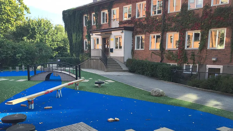 Röd tegelbyggnad i två plan med grönska och lekytor med gungbräda och liten lekbro i förgrunden.