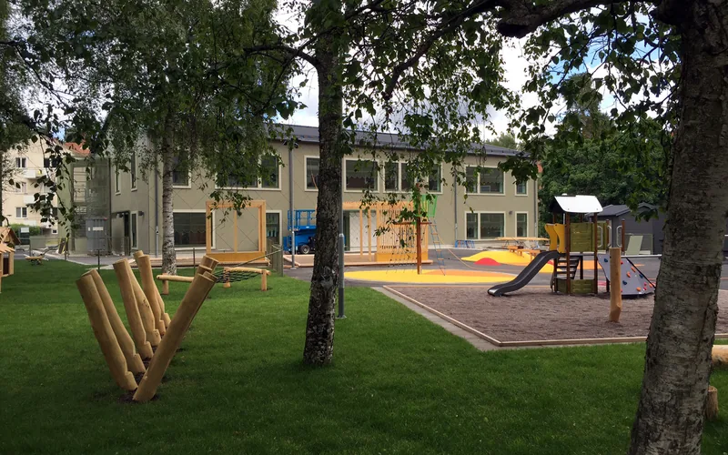 Förskolan i bakgrunden och gården med lekredskap och några björkar i förgrunden.
