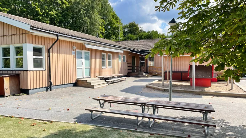 Förskolegård med sandlåda och lekhus samt gräsmatta, träd, bänkar och asfalt.