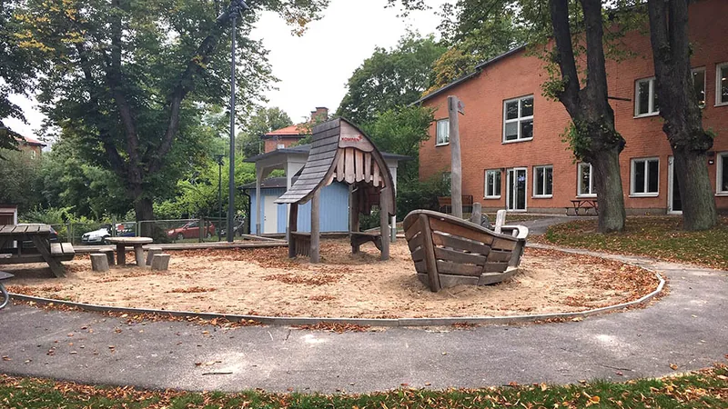 Röd tegelbyggnad i två plan med grönska och lekplats med sandlåda, sittplatser och lekhus i förgrunden.