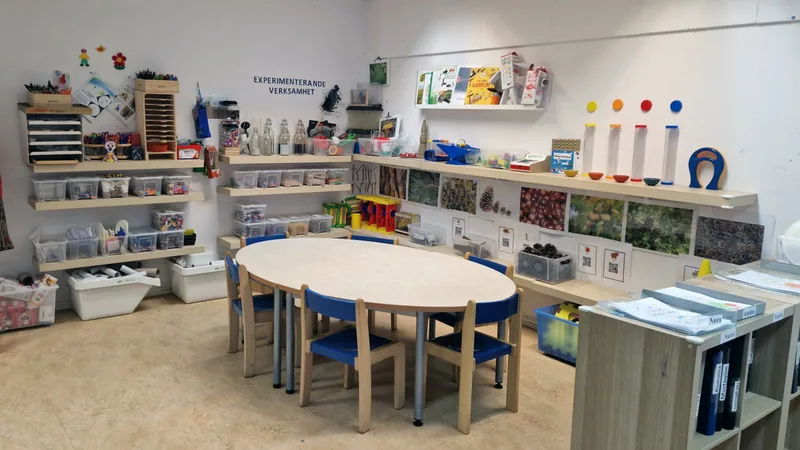 Ett lekrum med bord och stolar och hyllor fyllda med lekmaterial. På väggen står det skrivet "Experimenterande verksamhet".
