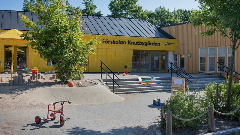 Förskolegård med träd, buskar och sandlåda med parasoll. I övrigt asfalt med trappor, trehjulingar, bollar och andra leksaker.