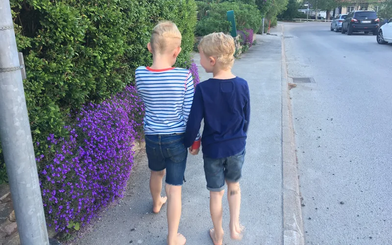 Två sommarklädda barn går och pratar på trottoaren. Blå blommor och gröna buskar.