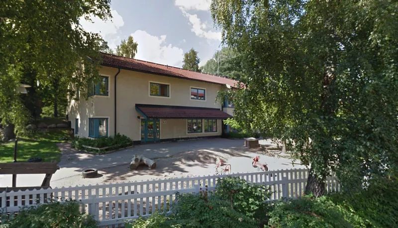 Förskolans fasad med en grå byggnad, lekplats och vita staket.
