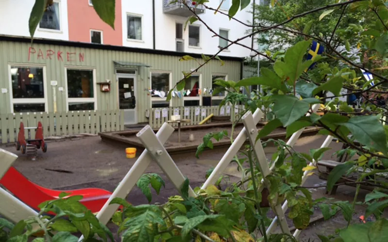 Utemiljö med lekytor, sandlåda och rutschkana. Ljusgrön byggnad i bakgrunden och spaljé med klätterväxt i förgrunden.