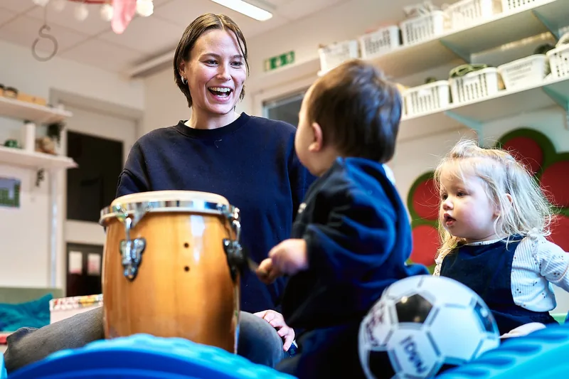 En vuxen person sitter och spelar på en kongatromma med två barn, ett av barnen håller i en fotboll. De ser glada ut i en lekmiljö.