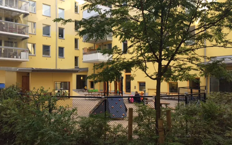 Utemiljö med lekytor, sandlådor och klätterställning med buskar och träd i förgrunden. Gult flerbostadshus i bakgrunden.