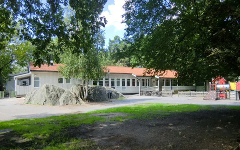 Vit enplansbyggnad med gård med grönska, litet berg och klätterställning.