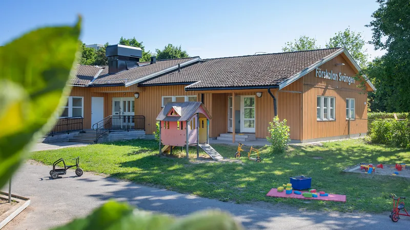 Förskolegård med gräs, buskar, träd och asfalt, samt klätterborg, sandlådor, trehjulingar och andra leksaker.