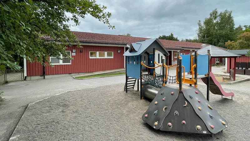 Förskolegård med klätterställning, sandlåda, rutschkana och träd.