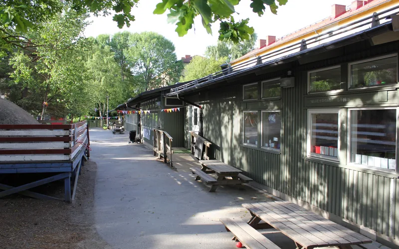 På högersida ser man förskolans gröna fasad med klassiska parkbänkar utanför. På vänstersida skymtar man förskolans gård.