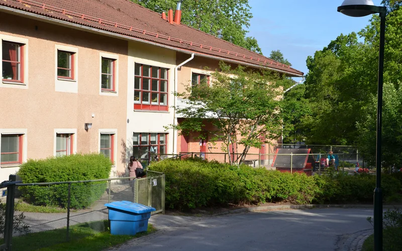 Husets entré omringad av grönska, bit av gården med lekytor i bakgrunden.