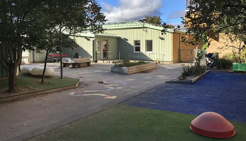 Ljusgrön byggnad i ett plan med lekytor, odlingar och växtlighet i förgrunden.