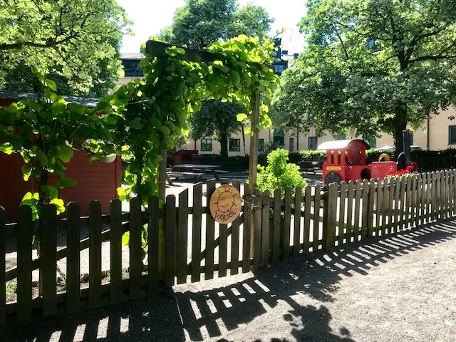 ingång till förskolans gård, grind, grönskande träd och buskar