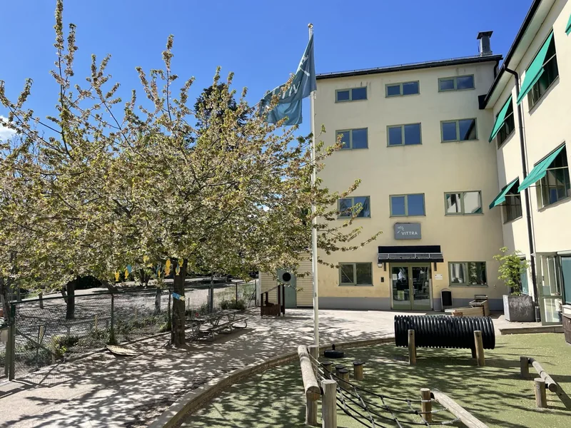 En ljusgul fyravåningsbyggnad med en gård framför som har lekverktyg och en flaggstång.